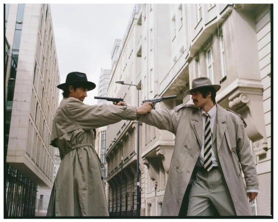 Two men in trench coats and fedoras standing in an urban street holding silenced pistols pointed at each other
