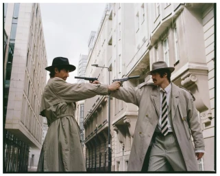Two men in trench coats and fedoras standing in an urban street holding silenced pistols pointed at each other