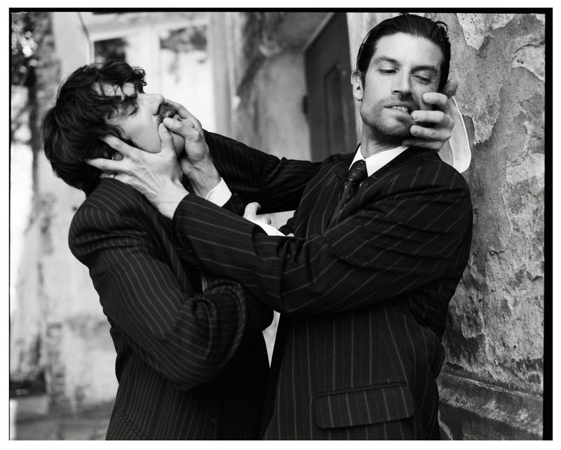 Black and white editorial portrait of two men in pinstriped suits interacting dramatically outdoors.
