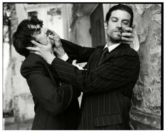 Black and white editorial portrait of two men in pinstriped suits interacting dramatically outdoors.