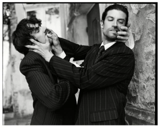 Black and white editorial portrait of two men in pinstriped suits interacting dramatically outdoors.