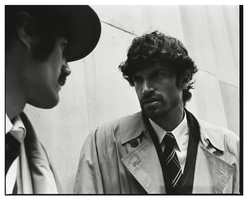 Black and white portrait of a man in a trench coat and tie conversing with another man in a hat.