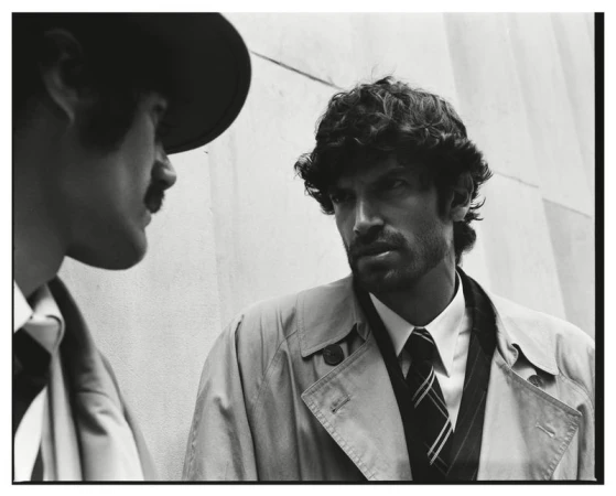 Black and white portrait of a man in a trench coat and tie conversing with another man in a hat.