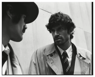 Black and white portrait of a man in a trench coat and tie conversing with another man in a hat.