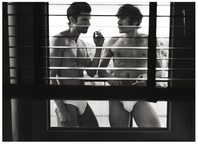Two shirtless men in white underwear standing on a balcony viewed through horizontal window blinds in a black and white editorial photograph.