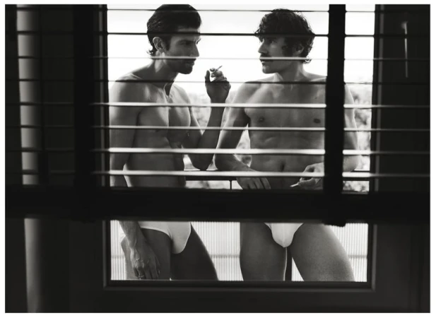 Two shirtless men in white underwear standing on a balcony viewed through horizontal window blinds in a black and white editorial photograph.