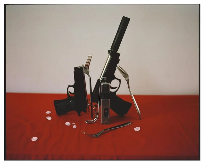 Two black handguns with silencers arranged with stainless steel forks and petals on a red table.