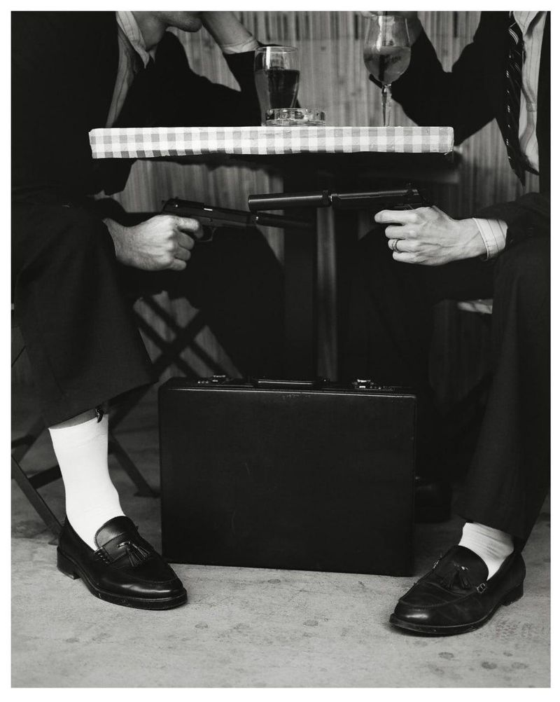 Two men in suits seated at a table pointing guns at each other with a briefcase below