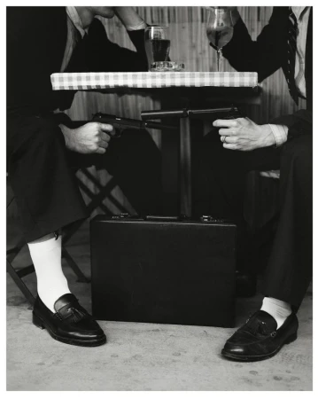 Two men in suits seated at a table pointing guns at each other with a briefcase below