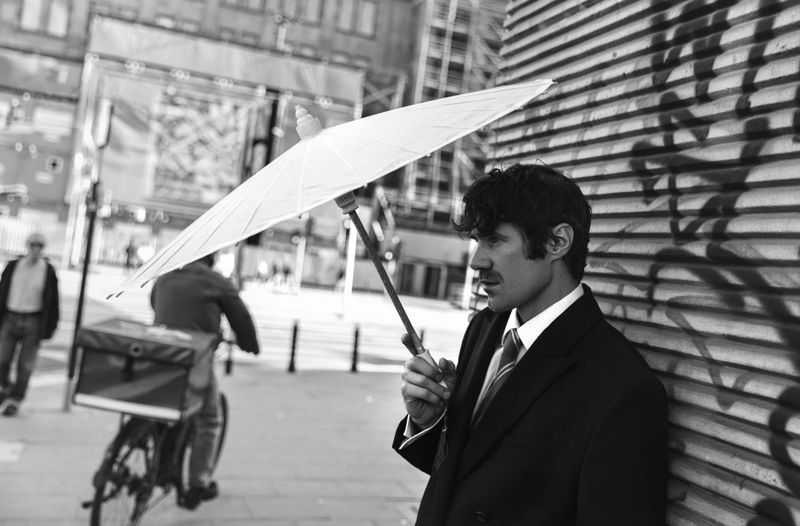 Man in a dark suit holding a white parasol standing against a corrugated metal wall on a city street.