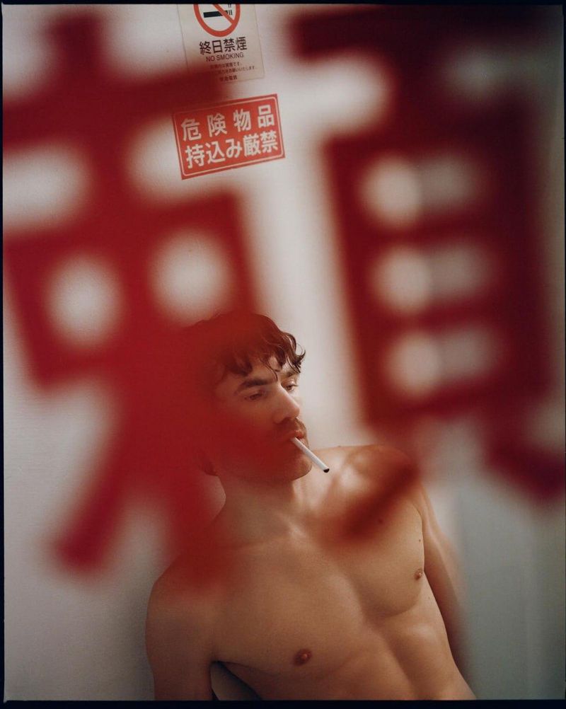 Shirtless man with a cigarette in his mouth framed behind red Japanese signage in an editorial portrait by Arthur Iskandarov.