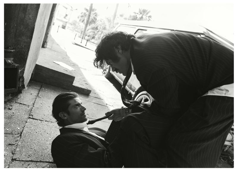 Black and white editorial photograph of two men in suits, one standing over the other on a city street.