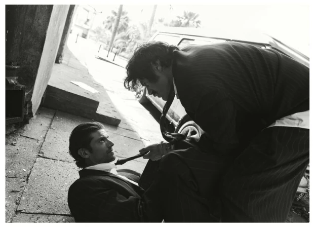 Black and white editorial photograph of two men in suits, one standing over the other on a city street.