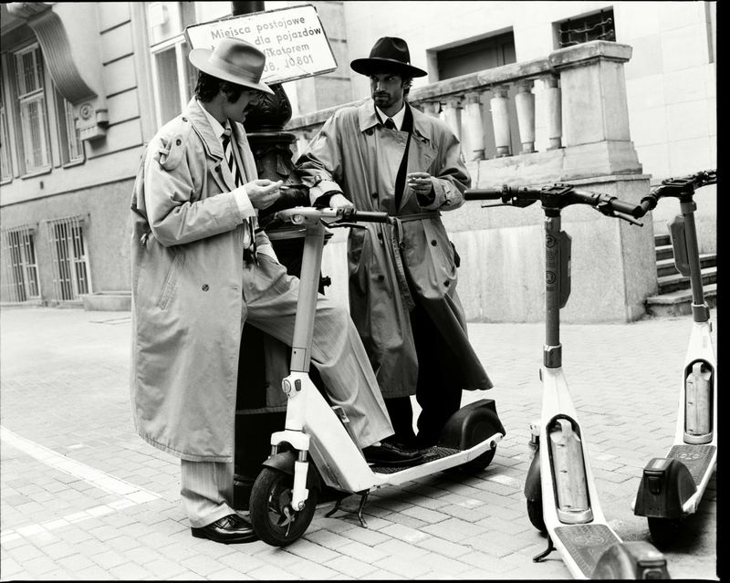 Two men in trench coats and fedoras standing on a city street beside electric scooters.