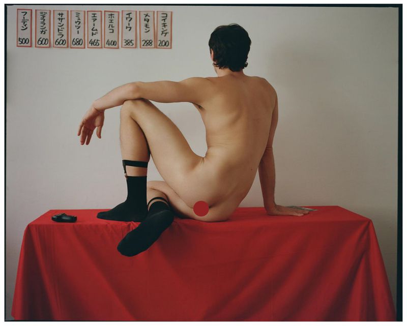 A man seated from behind on a red draped table wearing black socks against a white wall.