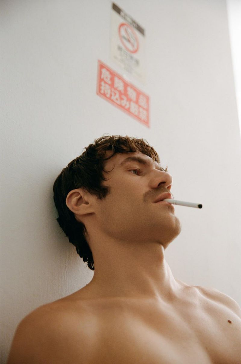 Shirtless male model with dark hair and mustache leaning against a white wall with a cigarette in his mouth.
