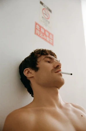 Shirtless male model with dark hair and mustache leaning against a white wall with a cigarette in his mouth.