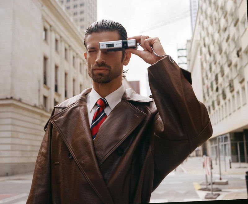 Model wearing a brown leather coat and striped tie holding a pocket camera to his eye outdoors on a city street.