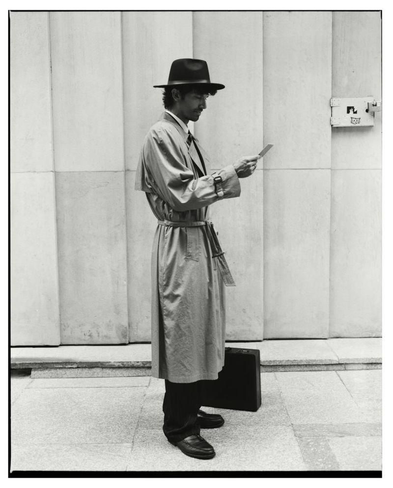 Man wearing a fedora and trench coat holding a small card outside a building for Numero Netherlands Trust editorial.