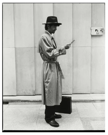 Man wearing a fedora and trench coat holding a small card outside a building for Numero Netherlands Trust editorial.