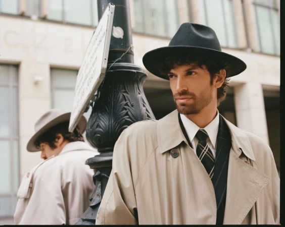Man wearing a black fedora and beige trench coat posing outdoors near a stone column for Numero Netherlands.
