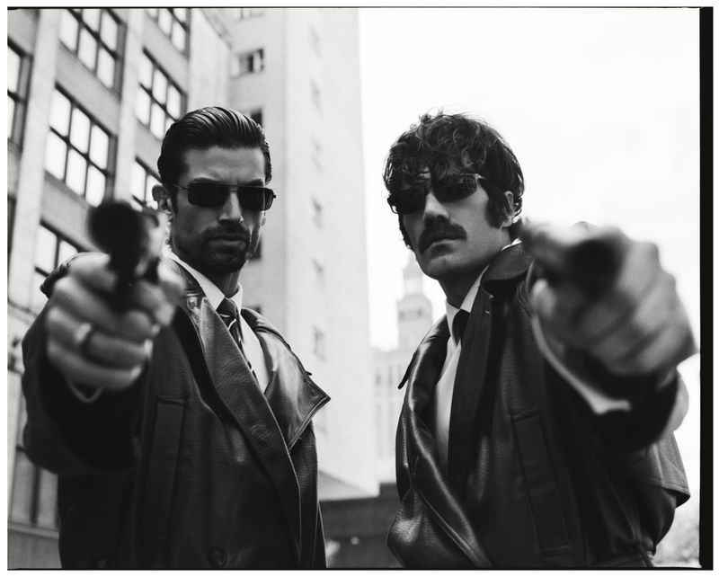 Two men in leather coats and dark sunglasses aiming pistols forward in a dramatic urban editorial photograph.