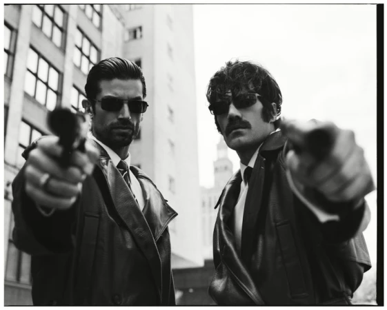 Two men in leather coats and dark sunglasses aiming pistols forward in a dramatic urban editorial photograph.