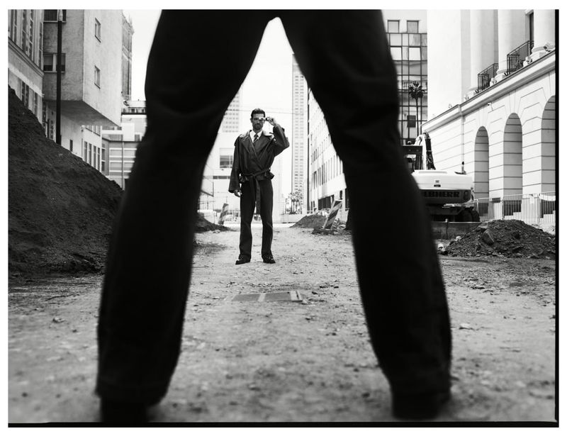 Low angle monochrome fashion editorial shot looking through a pair of legs at a man standing on a city street.
