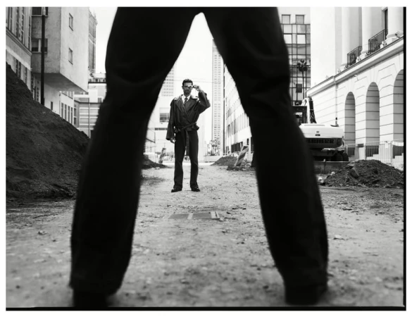 Low angle monochrome fashion editorial shot looking through a pair of legs at a man standing on a city street.