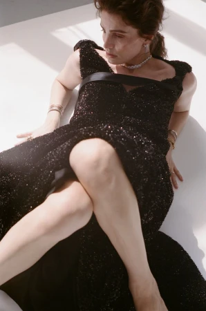 Model wearing a sequined black dress and diamond jewelry reclining against a bright white studio backdrop