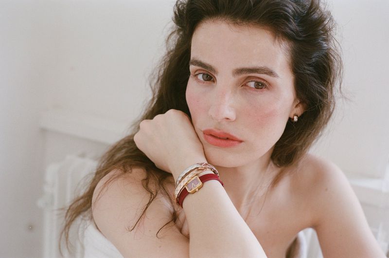 Close-up portrait of a woman resting her chin on her hand, wearing layered bracelets and a red-strap wristwatch against a neutral background.