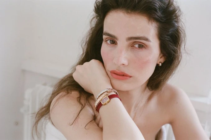 Close-up portrait of a woman resting her chin on her hand, wearing layered bracelets and a red-strap wristwatch against a neutral background.