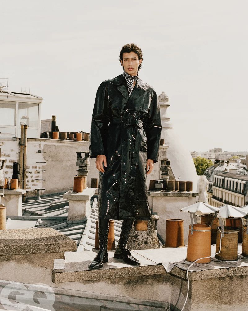 Full-length portrait of a male model wearing a glossy black leather coat standing on a city rooftop amid chimneys.