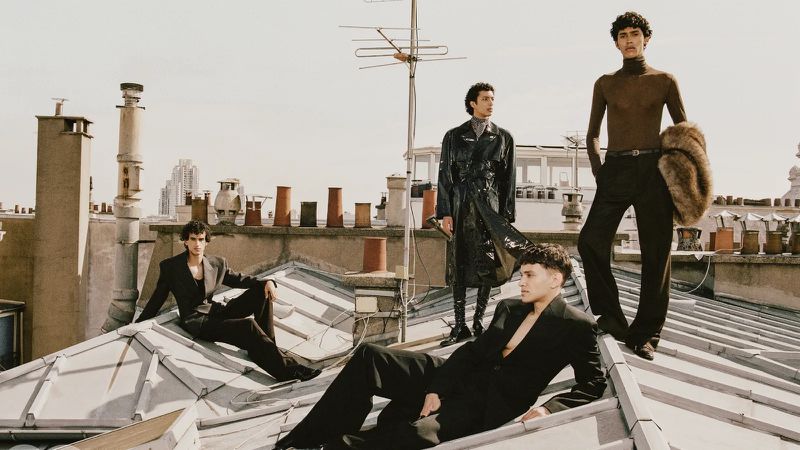 Four male models posing in designer fashion on a zinc rooftop with chimneys against an urban skyline background