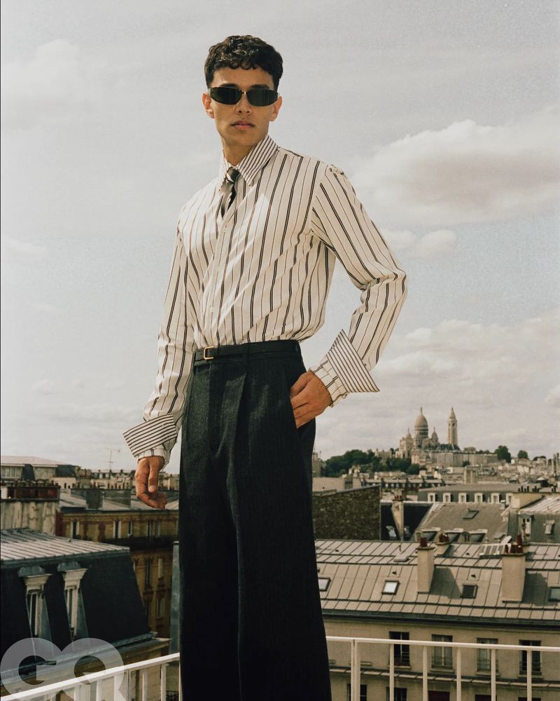 Model wearing dark sunglasses, striped shirt, and black trousers on a rooftop overlooking a cityscape under an overcast sky.