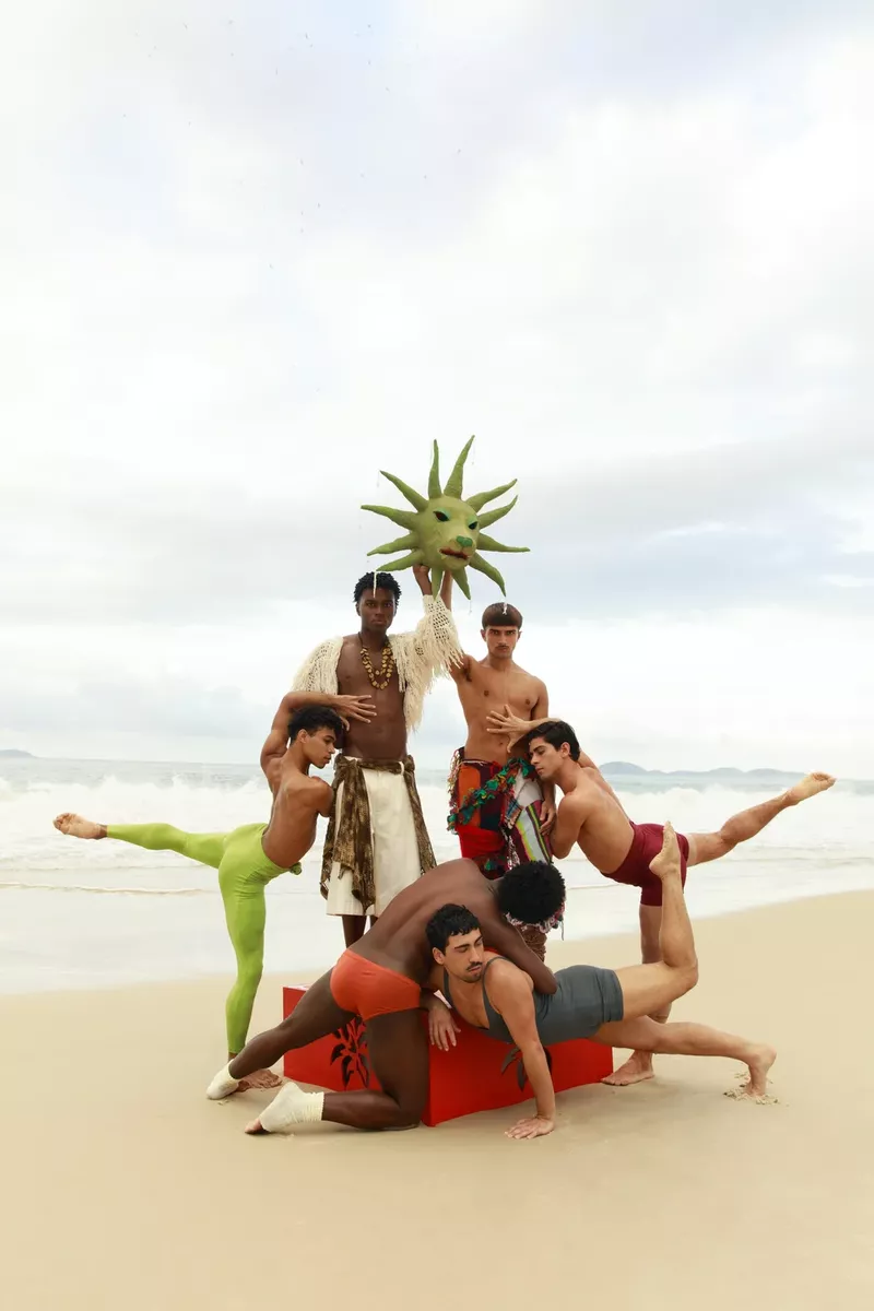 Group of six male dancers posing dynamically on a sandy beach under an overcast sky holding a starburst mask
