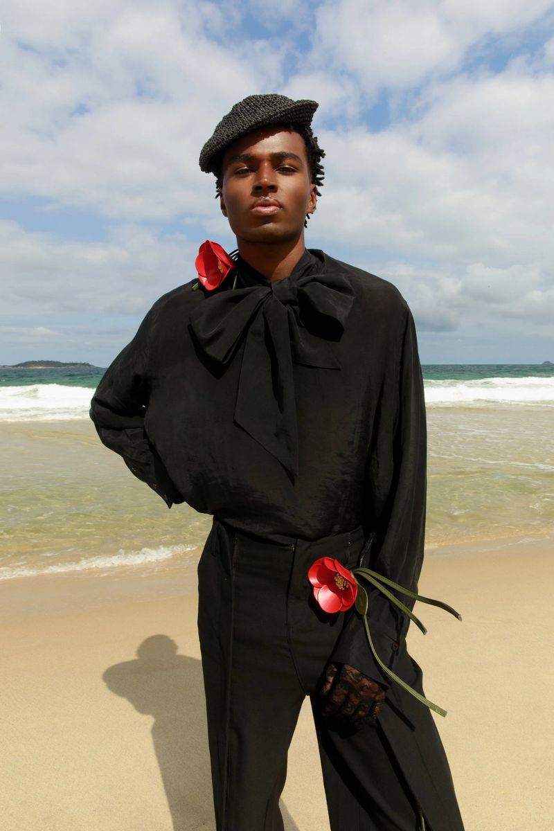 Black man wearing black silk blouse and trousers with red flowers on sandy beach by ocean under clear sky
