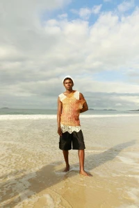 A man wearing a patterned sleeveless top and dark shorts stands on a sandy beach under a cloudy sky.