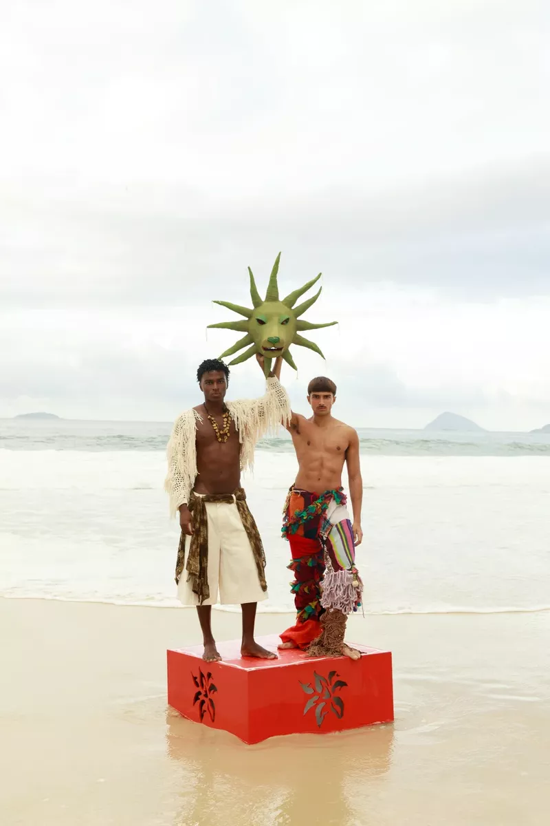 Two male models standing on a red box holding a green sun mask on a sandy beach for Numero Netherlands