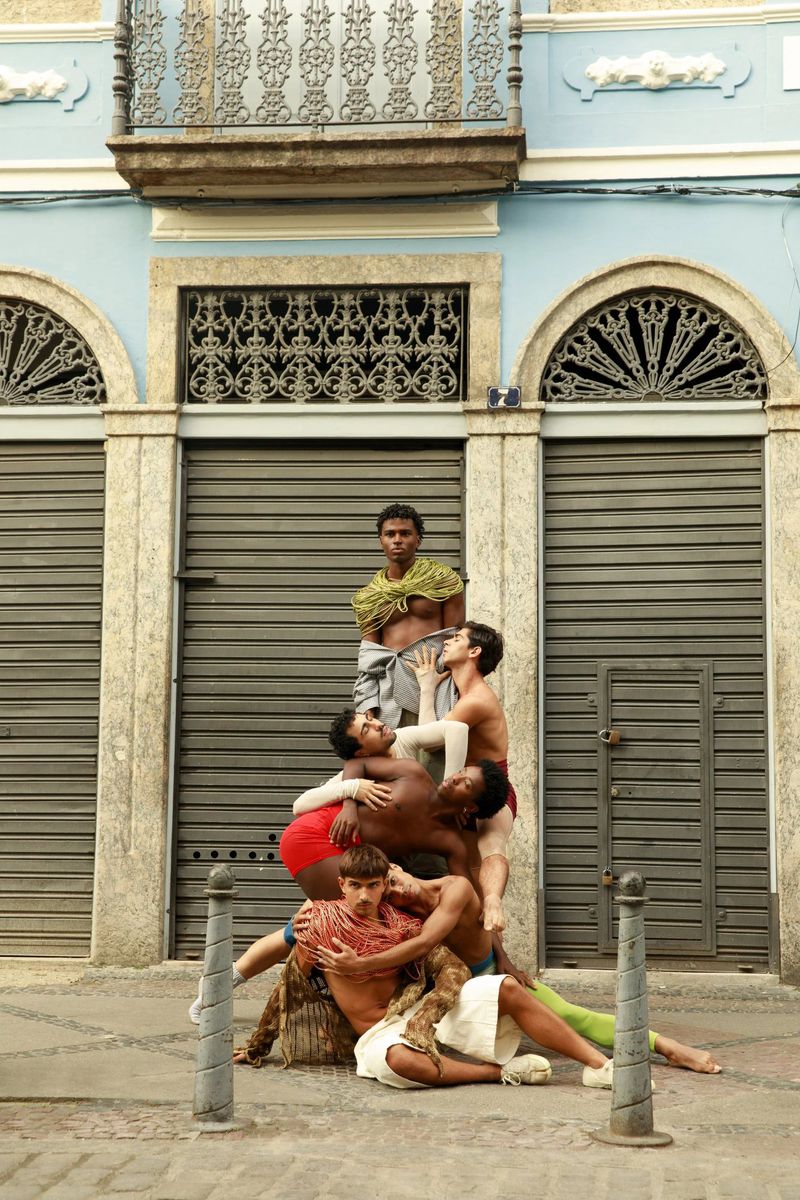 Group of six male models posed in a human pyramid on a cobblestone street in front of a blue building with closed metal shutters.