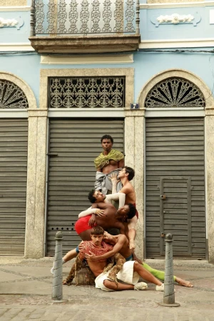 Group of six male models posed in a human pyramid on a cobblestone street in front of a blue building with closed metal shutters.