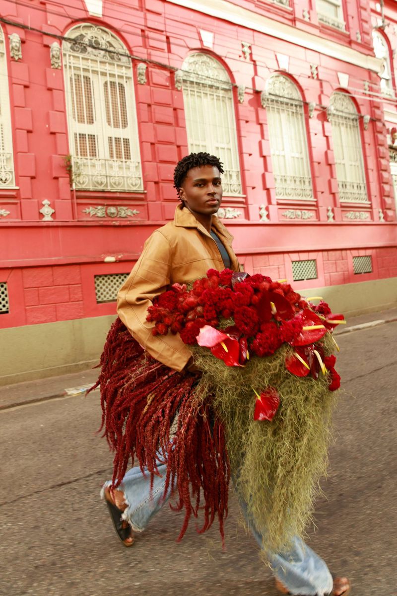 Black man holding a large arrangement of red flowers walking on a street past a red building.