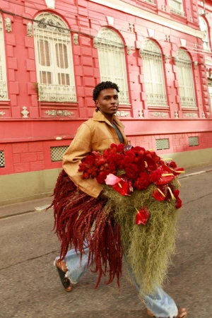 Black man holding a large arrangement of red flowers walking on a street past a red building.