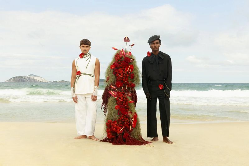 Two men standing on a sandy beach flanking an artistic floral sculpture with red flowers