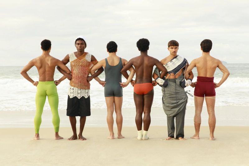 Six male models standing in a row with backs to the camera on a sandy beach with the ocean behind them.