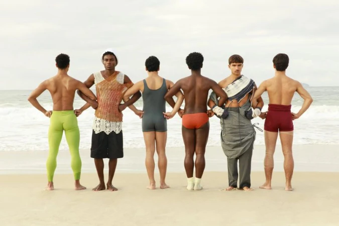 Six male models standing in a row with backs to the camera on a sandy beach with the ocean behind them.