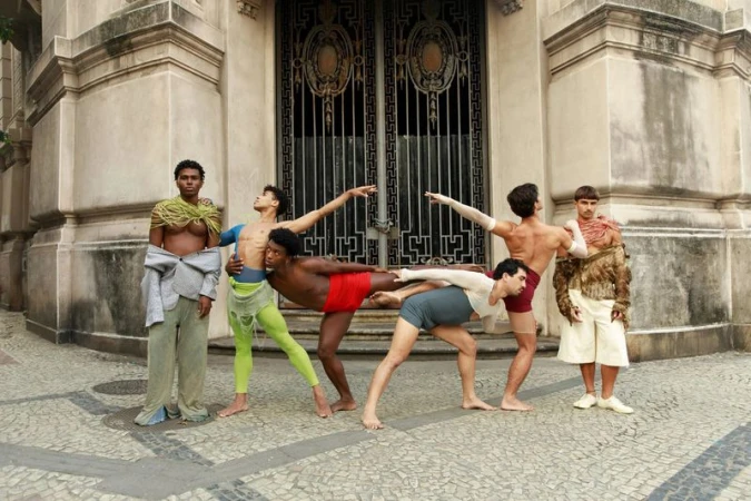 Group of six male dancers posing dynamically in front of a grand architectural doorway on a cobblestone street.