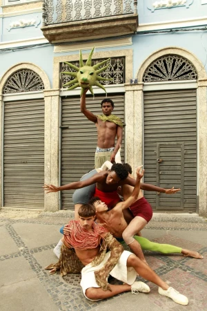 Group of men forming a sculptural human pyramid in front of a building facade holding a green sun mask