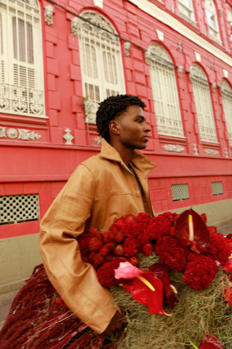 A young Black man wearing a tan leather jacket holding a large arrangement of red flowers against a vibrant pink building