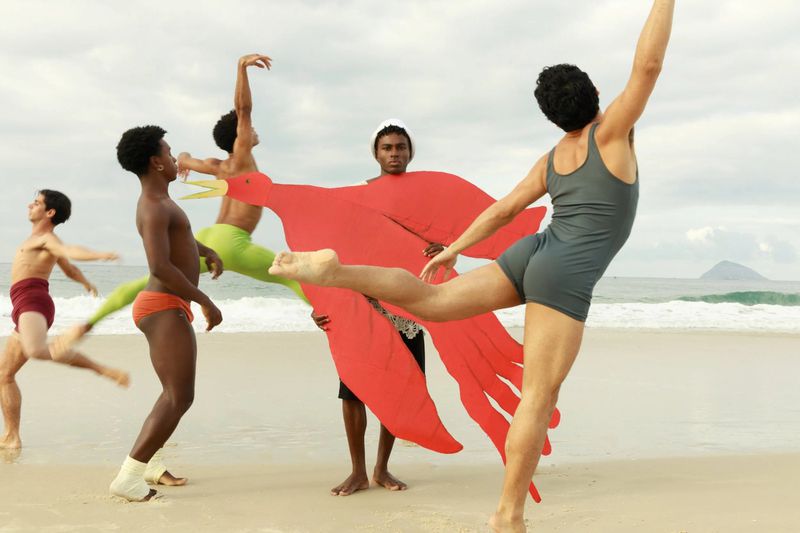 Group of male dancers on a sandy beach holding a large red paper cutout of a bird, captured in bright daylight.